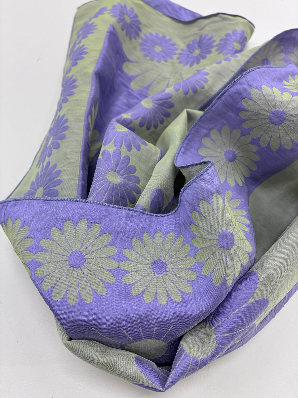 A lavender purple scarf with a light sage green daisy pattern, draped to show its woven texture and double-sided floral design.